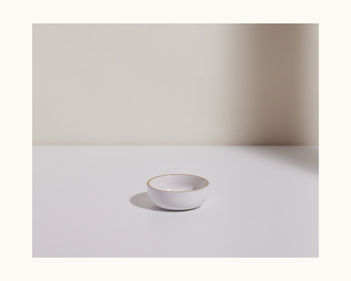 A Tiny Bowls scratch-resistant white ceramic bowl with a thin gold rim rests on a white surface, set against a light beige background.