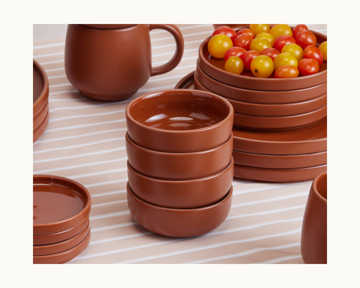 Tiny Bowls ceramic tableware, including stackable bowls, plates, and a mug, sits on a striped tablecloth with cherry tomatoes in the background. This scratch-resistant Tiny Bowls set brings both style and durability to your table.