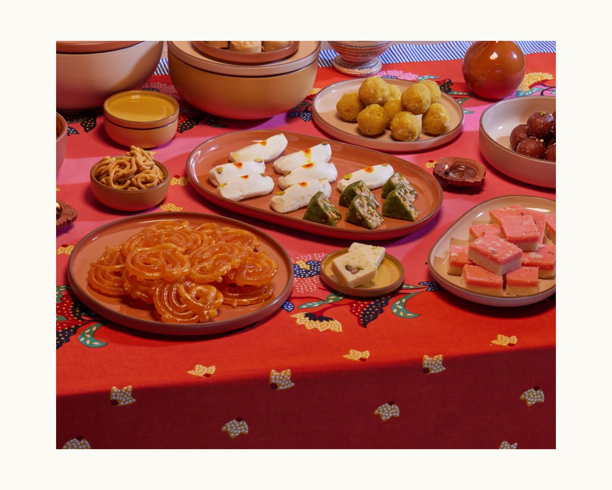 The Tiny Plates tableware set showcases Indian sweets like jalebi, barfi, peda, and rasgulla, plus savory snacks, all beautifully arranged on ceramic plates atop a red patterned tablecloth.