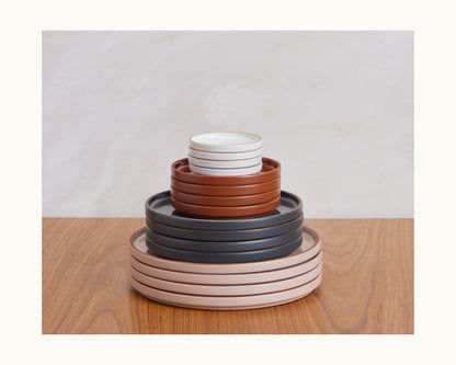 Tiny Plates ceramic tableware set, featuring plates and bowls in white, brown, gray, and beige—neatly stacked on a wooden surface with a light backdrop.