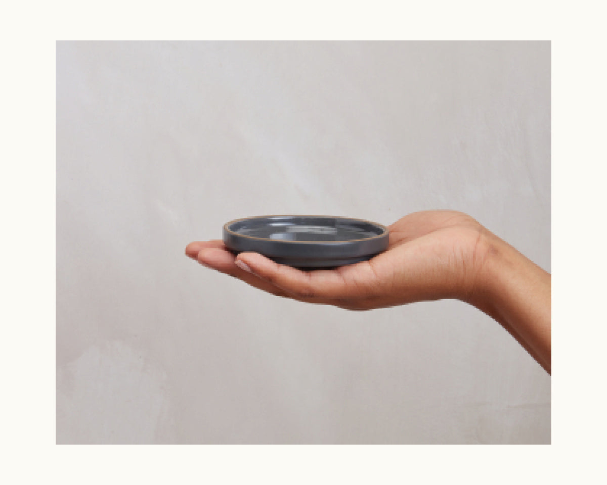A hand holds a black round plastic lid from Tiny Plates, perfect for covering snacks or modern tableware, set against a plain light background.
