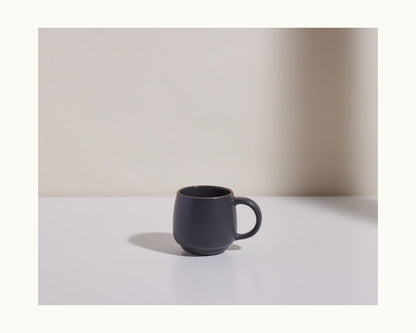 The Night + Day Mug, crafted from dark gray ceramic stoneware, rests on a white surface with a light gray background. Shadows to the left accentuate the mug’s generous capacity.