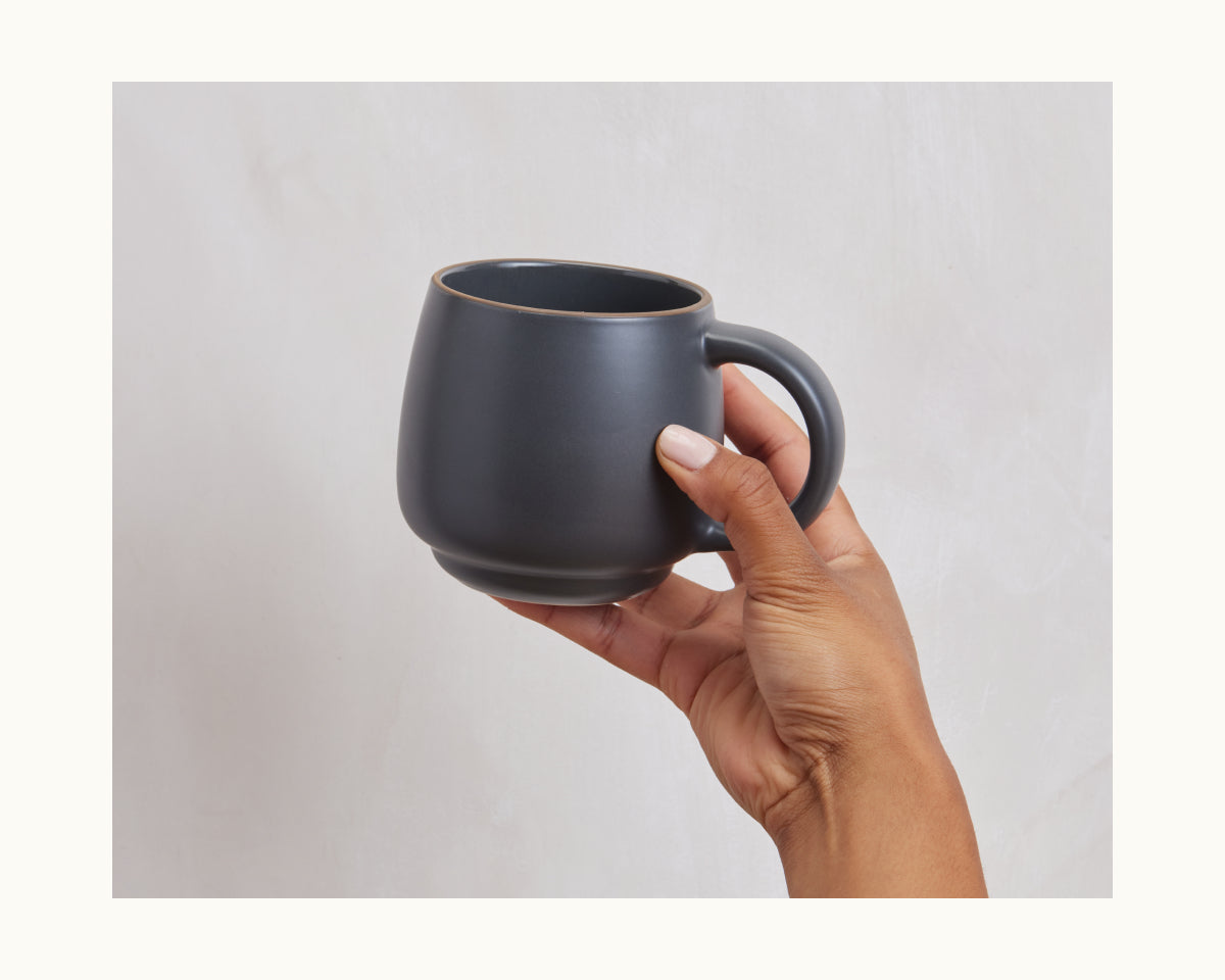 A hand holds a Night + Day Mug in matte dark gray ceramic stoneware with generous capacity, set against a light neutral background.