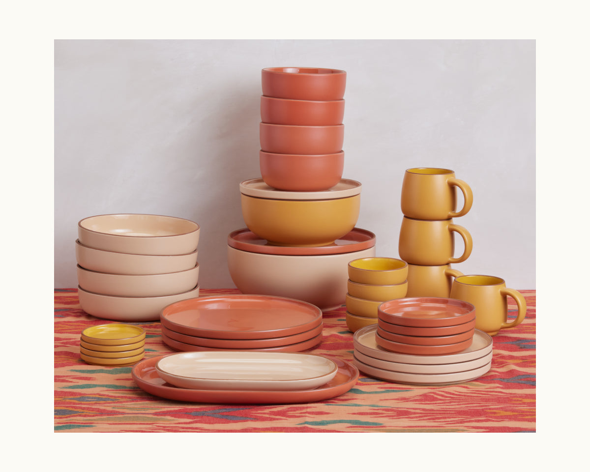 Night + Day Mugs in beige, terracotta, and mustard hues are stacked with matching bowls and plates on a vibrant red-orange patterned tablecloth.