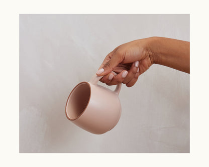 A hand with light pink nails holds a plain, pale pink Night + Day Mug made from ceramic stoneware with generous capacity, set against a light, textured background.
