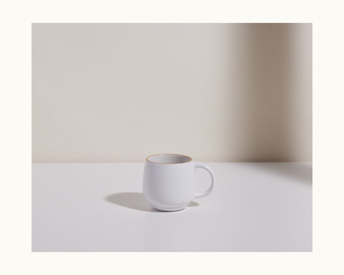 The Night + Day Mug, made from white ceramic stoneware with a handle, sits on a smooth white surface. Stackable in design, it provides ample capacity for your favorite beverages against a light background.