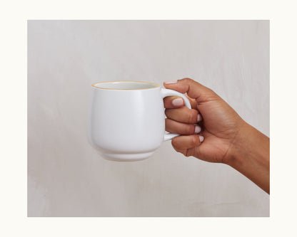 A hand holds the Night + Day Mug, a plain white ceramic stoneware mug with a generous capacity, against a light-colored background.