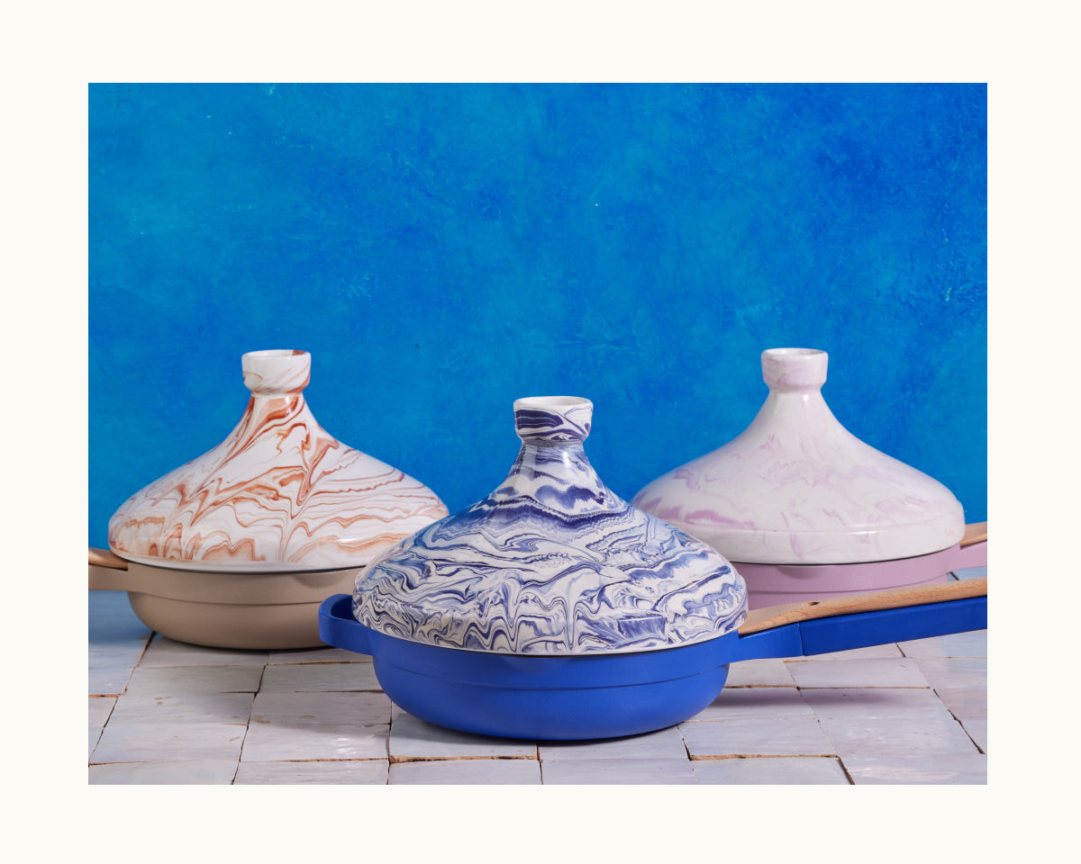 Three handcrafted Tagine pots with marbled orange, blue, and purple designs rest on a white tiled surface against a bright blue background—ideal for slow-cooked flavorful meals.