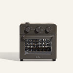 The AU - Wonder Oven is a dark gray countertop toaster oven with three top control dials and a front glass door featuring a diamond-patterned interior. "Our Place" appears at the bottom of the door. Doubles as an air fryer for extra versatility.