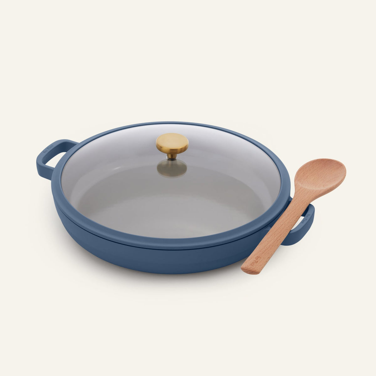 The Dual Handle Always Pan in blue, featuring a nonstick surface, glass lid, and gold knob, is placed next to a wooden spoon on a light background.