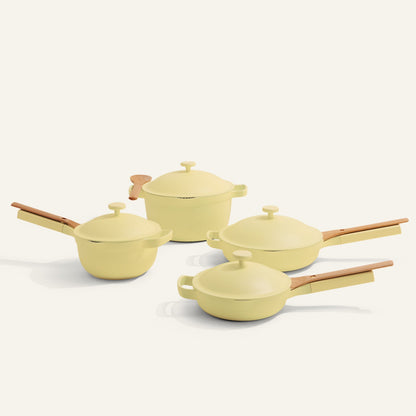 The Cookware Set includes four yellow pieces with wooden handles—a Dutch oven, a sauté pan, and two lidded saucepans—displayed on a light surface, in a style similar to the Always Pan and Perfect Pot.