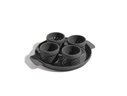 The Egg Poacher is a black silicone set with four perforated cups on a round tray with side handles, ideal for making poached eggs. Shown on a white background.
