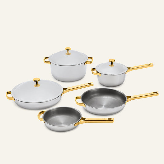The Titanium Pro Cookware Set+ 2.0 includes a large pot, a saucepan, two frying pans, and a lidded skillet, all featuring silver-toned metal bodies with gold-toned handles and lids.