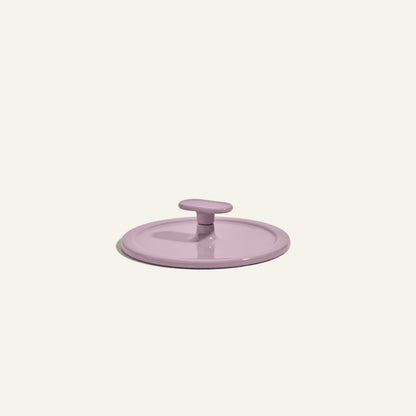 A round, light purple Grill Press with a central handle, crafted from cast iron for even heat distribution, displayed on a plain off-white background.