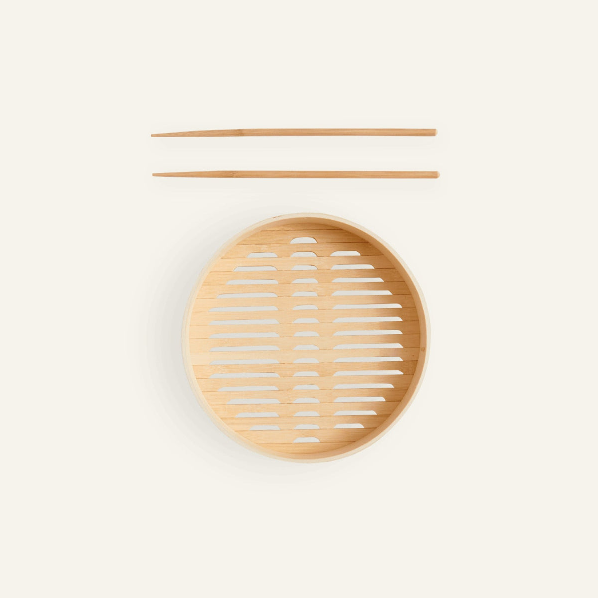 A pair of bamboo cooking chopsticks rests above the Spruce Steamer basket—a round steamer basket with a slotted base—set against a plain light background.