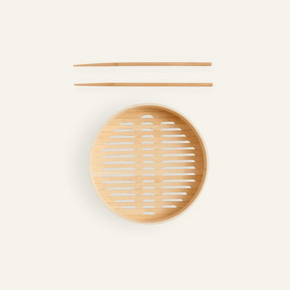 A pair of bamboo cooking chopsticks rests above the Spruce Steamer basket—a round steamer basket with a slotted base—set against a plain light background.